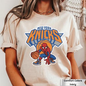 May include: An ivory t-shirt featuring a graphic of the New York Knicks logo with the text New York Knicks above a basketball and a cartoon Spider-Man character dribbling a basketball while wearing blue and orange athletic sneakers.