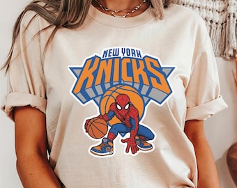Op de afbeelding: Een ivoorkleurig t-shirt met een afbeelding van het New York Knicks-logo met de tekst New York Knicks boven een basketbal en een cartoonfiguur van Spider-Man die een basketbal dribbelt en blauwe en oranje sportschoenen draagt.