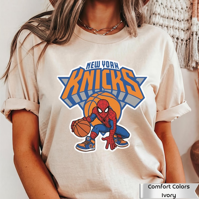 May include: An ivory t-shirt featuring a graphic of the New York Knicks logo with the text New York Knicks above a basketball and a cartoon Spider-Man character dribbling a basketball while wearing blue and orange athletic sneakers.