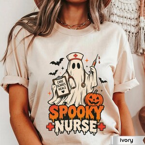May include: An ivory t-shirt featuring a graphic of a ghost dressed as a nurse with a stethoscope, a syringe, and a clipboard that reads CARE SCARES HEALS. Below the ghost is a jack-o'-lantern and the text SPOOKY NURSE with red medical crosses.