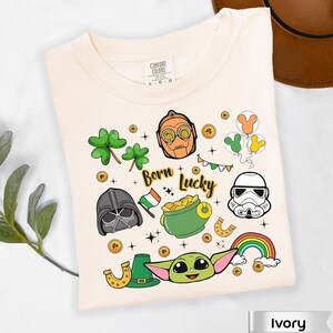 May include: Ivory-colored t-shirt with a graphic design featuring cartoon characters, shamrocks, a rainbow, and the text "Born Lucky." The design includes a pot of gold, a horseshoe, and a leprechaun hat.