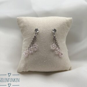 Könnte beinhalten: Hängeohrringe mit einer silbernen Kette und einer Ansammlung kleiner, hellrosa Perlen. Jeder Ohrring hat oben einen klaren, runden Kristall. Die Ohrringe werden auf einem kleinen, quadratischen, beigen Kissen präsentiert. Der Text "SEELENFUNKEN" ist sichtbar.