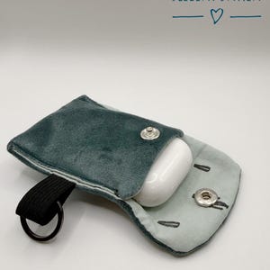 Könnte beinhalten: Eine tealfarbene Samttasche mit schwarzem Riemen und silbernem Ring, geöffnet, um ein weißes Kopfhörer-Etui zu enthüllen. Die Tasche hat einen Druckknopfverschluss. Der Text "SEELENFUNKEN" ist in Schreibschrift mit zwei Herzsymbolen geschrieben.