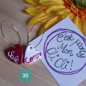 Peut inclure: Boucles d'oreilles en forme de cœur avec des cerceaux dorés. Une boucle d'oreille est rouge avec le mot "Sale" en blanc, l'autre est blanche avec "Comme" en rouge. Une carte avec du texte français et une fleur jaune en arrière-plan.