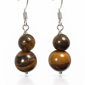 May include: A pair of drop earrings with two brown tiger's eye beads. The top bead is a darker brown, the bottom bead is lighter with dark brown stripes. The earrings have silver-coloured hooks and measure approximately 2 cm in length.