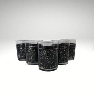 May include: A set of five cylindrical drinking glasses featuring a black exterior with a white marble-like vein pattern and a thin silver rim at the top edge. The glasses are arranged in a cluster, showcasing the consistent dark design.