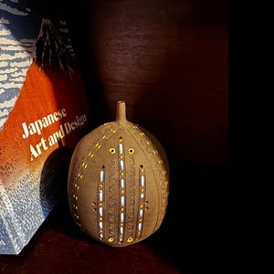 May include: A round, brown ceramic vessel with a narrow neck, featuring vertical rows of intricate, carved patterns inlaid with white and yellow accents. The object is positioned next to a book titled Japanese Art and Design.