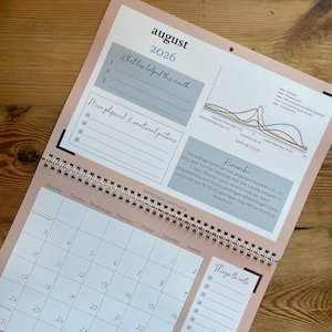 May include: A light pink and white spiral-bound calendar open to August 2026. It features a monthly calendar, note sections, and a graph. The calendar is displayed on a wooden surface.