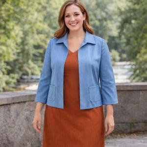 May include: A woman wearing a light blue jacket with three-quarter sleeves and two front pockets over a rust-colored dress. The jacket has a classic collar and a tailored fit. The background is a blurred outdoor scene with greenery.