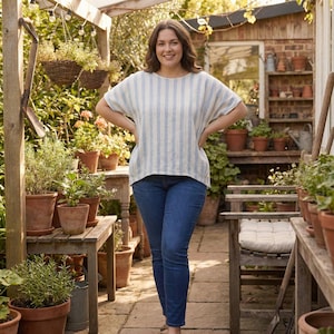 Puede incluir: Una mujer con una blusa a rayas azul claro y blanca y vaqueros azules posa en un jardín. La blusa tiene cuello redondo y mangas cortas. El jardín incluye plantas en macetas, muebles de madera y un edificio de ladrillo.