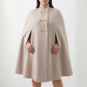 Puede incluir: Una capa beige con cuello alto y botones dorados. La capa tiene un diseño cruzado y una silueta fluida. La prenda presenta costuras verticales y aberturas para los brazos.