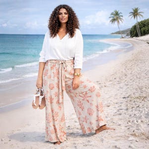 May include: A woman wearing a white blouse and beige wide-leg pants with a seashell and coral print. She is holding sandals and standing on a sandy beach with the ocean in the background. The pants have a matching tie belt.