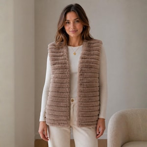 May include: A brown faux fur vest with a ribbed design. The vest is sleeveless and has a button closure. The model is wearing a white long-sleeve shirt and cream-colored pants. A gold necklace is visible.