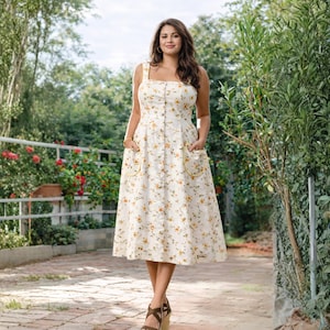 May include: A cream-colored sundress with a yellow floral pattern. The dress features a button-down front, square neckline, and two front pockets with yellow trim. The dress has a midi length and is worn with brown wedge sandals.