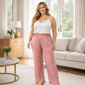May include: A woman wearing a white camisole with lace trim and pink pajama pants. The pants have a gold trim at the hem. The woman is standing in a well-lit living room with a sofa and a round coffee table.