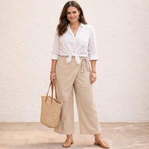 May include: A woman in a white shirt knotted at the waist, with beige wrap trousers. She carries a woven tote bag with leather handles and wears sandals. The outfit is casual and suitable for warm weather.