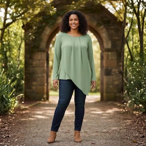 May include: A woman wearing a sage green long-sleeve top with an asymmetrical hemline and dark blue jeans. The top has a draped design. She is wearing brown ankle boots. The background features a stone archway and lush greenery.
