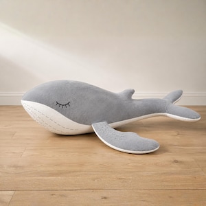 May include: A large, plush grey whale stuffed animal with a white belly and fins. The whale has a stitched-on closed eye and is lying on a wooden floor. A soft, cuddly toy for children.