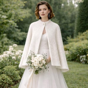 May include: A bride in a white wedding dress and a textured, off-white cape. The cape is fastened with a single button at the neck. She holds a bouquet of white roses and greenery. The background is a blurred garden setting.