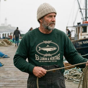 Peut inclure: Un homme porte un sweat-shirt vert avec un motif de poisson et le texte "SEA LABOR & HERITAGE THE OLD MAN AND THE SEA" sur un quai en bois. Un bateau de pêche est en arrière-plan, avec le nom "THE NANCY ELLEN" sur le côté.