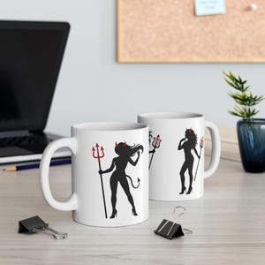 Peut inclure: Deux mugs en céramique blanche avec une silhouette noire d'une femme diable tenant un trident rouge. La femme diable a des cornes et une queue. Les mugs sont sur une surface en bois de couleur claire.