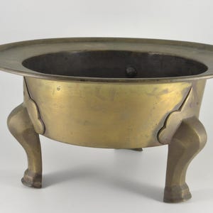 Puede incluir: Un cuenco de latón de estilo antiguo con un borde ancho y plano y tres patas curvas. El cuenco tiene una abertura circular y recortes decorativos donde las patas se unen al cuerpo. El metal tiene un aspecto ligeramente envejecido.