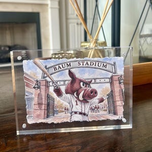 May include: A clear acrylic block frame containing a watercolor illustration of a mascot resembling a wild boar wearing a baseball uniform with the number 1, holding a bat in front of a brick archway labeled BAUM STADIUM with iron gates.