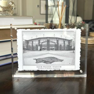 May include: A black and white print of Baum-Walker Stadium in a clear acrylic frame. The stadium is depicted with architectural detail, and the University of Arkansas Razorback logo is at the bottom. The text "Baum-Walker Stadium" and "University of Arkansas - Fayetteville" are visible.