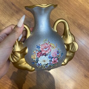 May include: A decorative vase with a silver body and gold accents. The vase features two elephant head handles and a floral design in the center. The vase has a ruffled top.