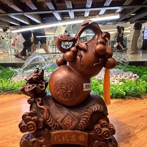May include: A carved wooden sculpture featuring a gourd shape with intricate details. The sculpture has a dark brown finish and includes a small, detailed figure on a base with Chinese characters. A yellow tassel hangs from the gourd.