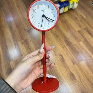 May include: A red desk clock with a round face and a matching base. The clock face is white with black numerals and hands. The clock is approximately 6 inches tall.