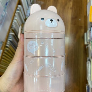 May include: A pink, three-tiered lunch container with a bear-shaped lid. The container has a cartoon bear face on top, with the words "Just Smile" and carrot and paw print designs. The bottom of the container has the words "Moshi Moshi".