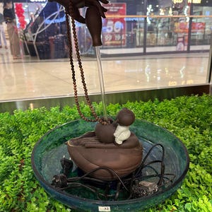 May include: A brown water fountain with a glass bowl and a small statue of a baby Buddha. The fountain has a hand-shaped element holding a string of beads. The bowl is surrounded by green foliage.