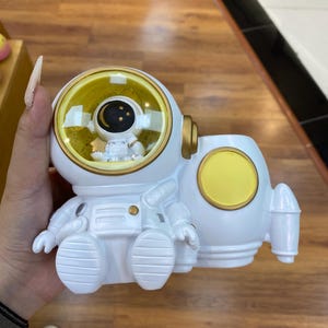 May include: White astronaut figurine with a yellow visor and accents. The astronaut is seated, with a rocket-shaped base. The visor reveals a small astronaut figure and a crescent moon.
