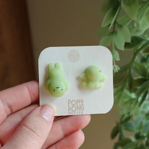 May include: A pair of pale green, translucent stud earrings shaped like small, cartoonish figures with simple black dot eyes and mouths. The earrings are attached to a square, off-white display card featuring the text PONG PONG PETITE.