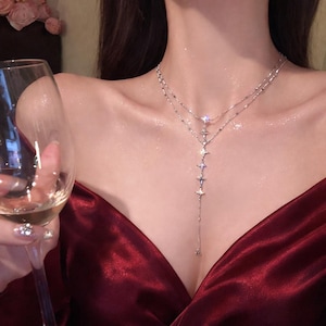 Könnte beinhalten: Eine silberfarbene, mehrschichtige Halskette mit sternförmigen Anhängern. Die Halskette hat ein filigranes Kettendesign mit mehreren Stern-Anhängern, die nach unten fallen. Der Schmuck wird vor einem tiefroten Stoff präsentiert.