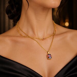 Puede incluir: Un collar de capas en tono dorado con dos cadenas. La cadena más corta es de estilo eslabones tipo clip con pequeñas piedras rojas incrustadas. La cadena más larga presenta un colgante rectangular con una gran piedra roja facetada.