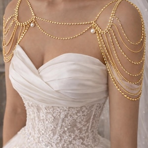 May include: A gold shoulder necklace featuring multiple strands of fine chains and tiny beads. The necklace is embellished with small pearls and is worn over a white wedding dress with a detailed lace bodice.