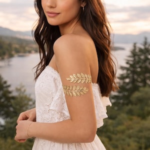 May include: Gold-colored leaf arm cuffs. The arm cuffs are made of metal and are designed to be worn on the upper arm. The leaves are detailed and arranged in a symmetrical pattern. The cuffs are shown on a person wearing a white dress.