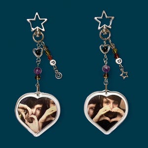 May include: Two heart-shaped keychains with a photo of two people making a heart shape with their hands. Each keychain has a silver star and heart charm, with colorful beads and the letters "P" and "D".