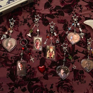 Puede incluir: Una colección de llaveros con imágenes de retratos en forma de corazón y rectangular. Cada llavero presenta cuentas decorativas, dijes y detalles en forma de estrella. También hay una pequeña botella de vidrio con un corcho y un objeto en forma de media luna.