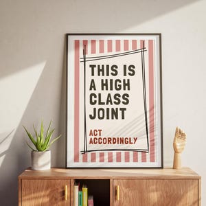 May include: Framed poster with the text "THIS IS A HIGH CLASS JOINT ACT ACCORDINGLY" in black and red. The background has a cream color with vertical pink stripes. The poster is in a black frame.