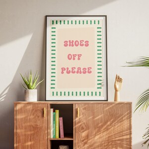 May include: A rectangular framed wall art print featuring a light beige background with a green dashed border and the pink text SHOES OFF PLEASE in a rounded font. The print is displayed in a dark frame, centered above a wooden cabinet.