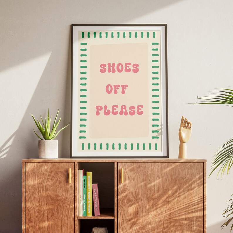 May include: A rectangular framed wall art print featuring a light beige background with a green dashed border and the pink text SHOES OFF PLEASE in a rounded font. The print is displayed in a dark frame, centered above a wooden cabinet.