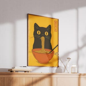 May include: A framed poster featuring a black cat eating noodles from a red bowl. The cat's silhouette is prominent against a yellow background. A black chopstick rests in the bowl. Japanese text is in the upper right corner.