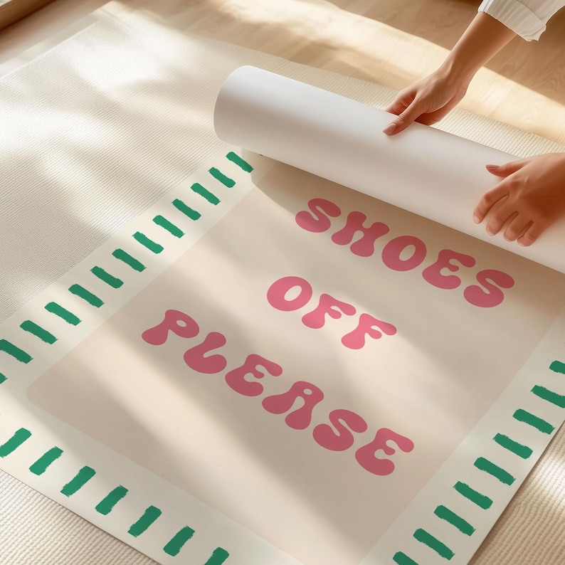 May include: A rectangular floor mat with a beige surface featuring the pink text SHOES OFF PLEASE in a rounded font. The mat has a border of short, green, brush-stroke style dashes along the left and right sides, and is partially rolled up.
