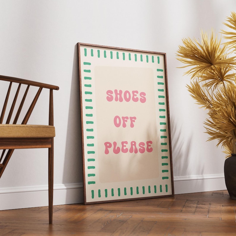 May include: A rectangular wall art print in a dark wood frame featuring a beige background with a green dashed border. The text SHOES OFF PLEASE is printed in the center in a pink, rounded, wavy font. The print is displayed in a vertical orientation.