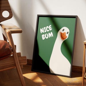 Op de afbeelding: Een ingelijste kunstdruk met een cartoon gans illustratie. De gans heeft een wit lichaam, een oranje snavel en zwarte ogen, tegen een groene achtergrond. Witte tekst luidt "NICE BUM". De lijst is zwart.