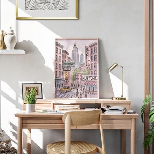 May include: Watercolor print of a New York City street scene, featuring the Empire State Building and a yellow taxi. The print includes the words "Broadway", "One Way", "Hotel", and "New York". The artwork is framed and displayed on a wooden desk.