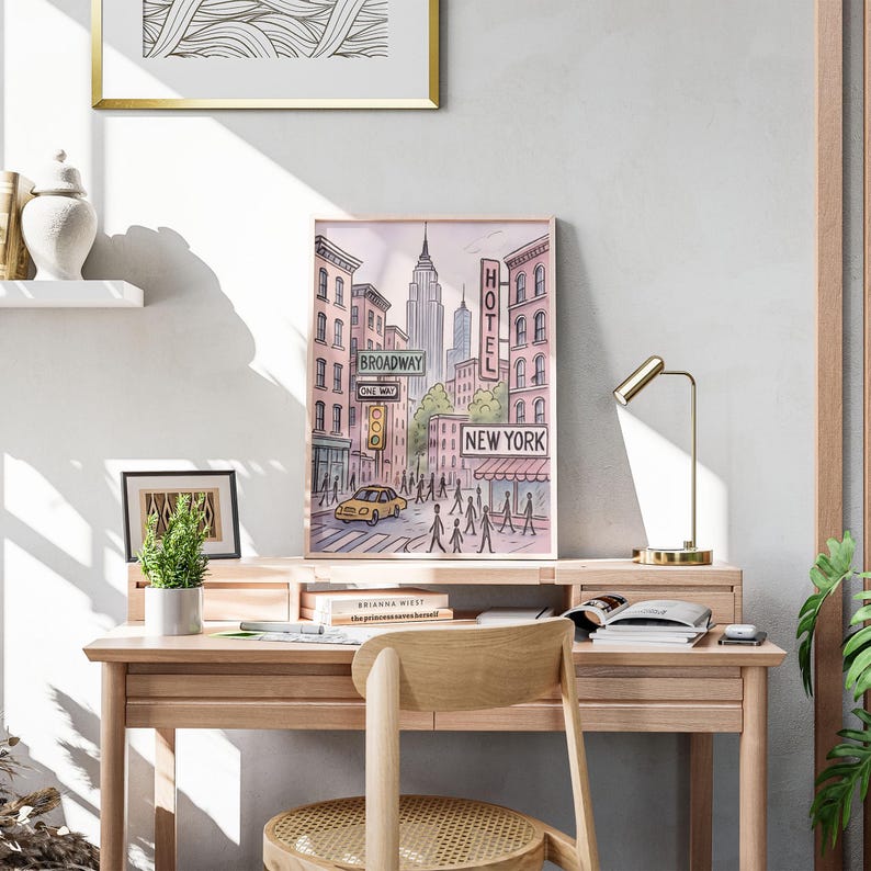 May include: Watercolor print of a New York City street scene, featuring the Empire State Building and a yellow taxi. The print includes the words "Broadway", "One Way", "Hotel", and "New York". The artwork is framed and displayed on a wooden desk.